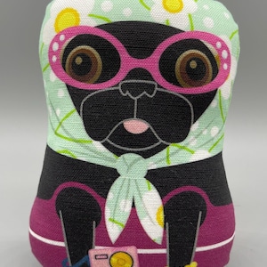 May include: A black pug wearing pink sunglasses and a green scarf with white flowers. The pug is sitting on a pink background with a small radio and a bracelet.
