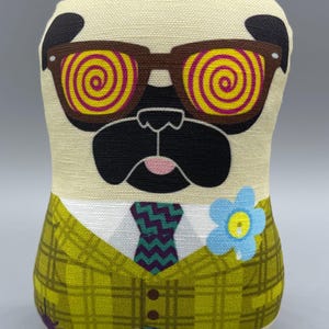 Puede incluir: Muñeco de peluche de un pug con cuerpo color crema, que lleva gafas de sol marrones con lentes en espiral amarillas y rosas. El pug viste una blazer a cuadros verdes, una corbata estampada y una flor azul.