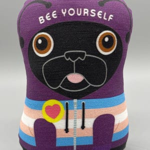 May include: A purple plush toy shaped like a pug with a black face and brown eyes. The toy is wearing a purple hoodie with a transgender pride flag design and a yellow heart on the chest. The text "BEE YOURSELF" is printed on the top of the toy's head.
