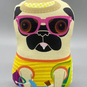 May include: A stuffed pug doll wearing pink sunglasses, a tan hat with a yellow chequered band, and a yellow striped shirt with colourful accents. The pug's face is black and tan with a pink nose.