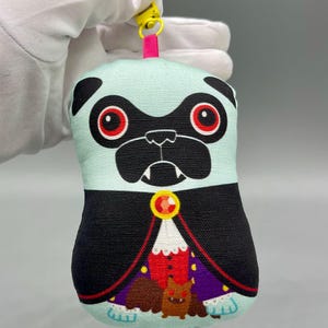 May include: A plush keychain featuring a light blue pug character with red eyes and black markings, dressed in a black vampire cape with a red and purple interior, a gold brooch, and a small brown bat graphic at the bottom with a yellow plastic clip.