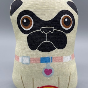 May include: A beige plush toy pug with black ears, a black nose, and brown eyes. The pug is wearing a pink and blue collar with a pink heart charm. A rainbow is printed on the bottom of the toy.