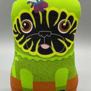 May include: A green plush toy shaped like a pug with a black face and a purple and yellow butterfly on its head. The pug is wearing a green sweater and orange trousers.