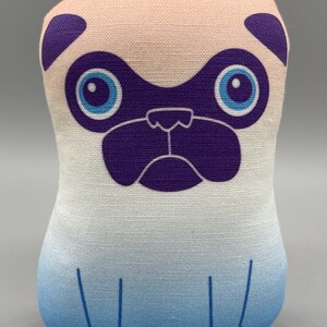 Peut inclure: Un jouet en peluche en forme de carlin avec un design dégradé rose et bleu. Le carlin a des oreilles violettes, un nez violet et des yeux bleus.