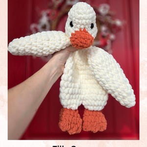May include: A white crocheted goose toy with orange feet and beak. The toy is being held by a hand in front of a red door. Tilly Goose Crochet Pattern by: LoopsAlot Crochet