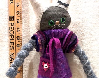 May include: A handmade cat doll with a dark grey head, white ears, and green eyes, wearing a purple jumper with a heart patch and white socks with black shoes. A wooden ruler placed next to the doll shows a height of approximately 46 centimetres.