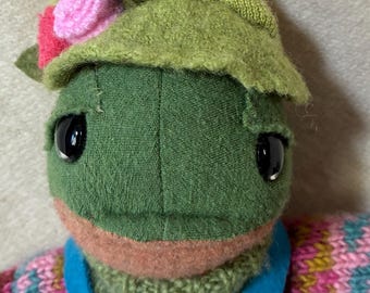 May include: A handmade green fabric doll with large black eyes, wearing a green hat with pink flowers, a blue jumpsuit with pink floral embroidery, and a multicoloured sweater. The doll has a whimsical, folk art style.