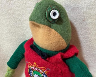 May include: A handmade frog doll with a green head, tan snout, and button eyes. It wears a red sweater with the "Slouth Golf Club" logo and green sleeves and legs. The doll is a whimsical, handcrafted item.
