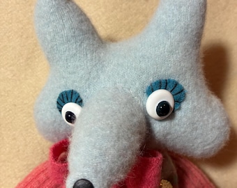 May include: A handmade fox-shaped plush toy. The fox has a light blue head and ears, with large, googly eyes and a black nose. It is dressed in a pink sweater over a blue sweater. A charming and whimsical creation.