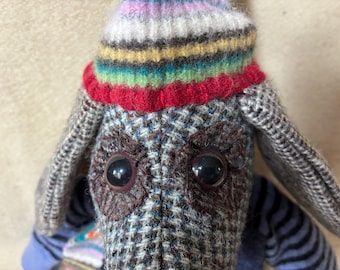 May include: A handcrafted fabric dog toy with large black eyes and a colourful striped hat. The dog's body is made of patterned fabric in shades of grey, blue, and brown. The hat features stripes of red, yellow, green, and blue.