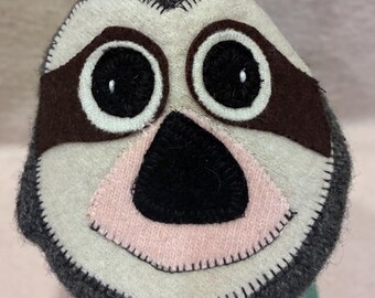 May include: A plush sloth face crafted from dark grey, cream, and light pink felted fabric. The design features large black circular eyes with white borders, dark brown eye patches, and a black triangular nose, all joined by visible black stitching.