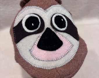 May include: A plush sloth head toy featuring a brown textured exterior with a cream-coloured face mask, large black circular eyes, dark brown eye patches, a black triangular nose, a light pink mouth area, and a small tuft of brown hair on top.