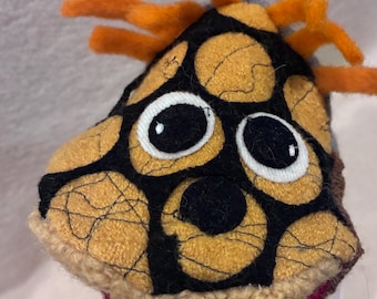 May include: A plush toy featuring a tan and black patterned top with stitched details, two large white and black eyes, and several bright orange felt strands extending from the crown. The base is a solid dark red fabric with a beige knitted trim.