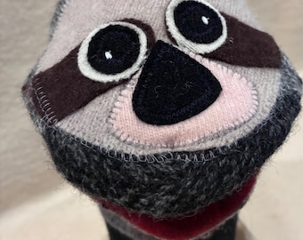 May include: A sloth-shaped puppet crafted from knit fabric in shades of grey, featuring dark brown felt eye patches, black embroidered eyes, a black felt nose, and a red fabric mouth. The puppet has a textured, multi-tonal grey knit exterior.