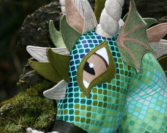 Könnte beinhalten: Ein Plüschdrachenspielzeug in Türkis-, Grün- und Brauntönen. Der Drache hat einen gemusterten Körper, große Augen und gebogene Hörner. Ein dunkelbrauner Riemen mit Knopfdetail umschließt den Hals. Der Drache befindet sich vor einem verschwommenen natürlichen Hintergrund.