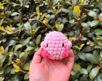 May include: A pink crocheted plush toy shaped like a blobfish with pink cheeks and black eyes.