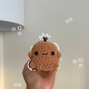 Może przedstawiać: Ręcznie robiona, pluszowa, brązowa lalka amigurumi z czarnymi oczami i wyszywanym uśmiechem. Lalka ma małe ramiona i detal w kształcie korony na górze. Lalka jest trzymana w dłoni na neutralnym tle z bazgrołami gwiazd.