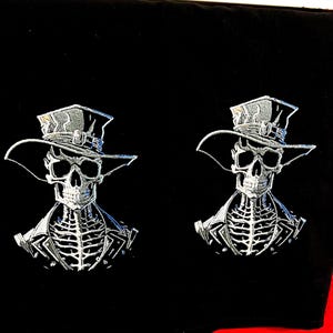May include: Two embroidered patches featuring a skeleton wearing a hat and a jacket. The skeleton is in a silver color against a black background.