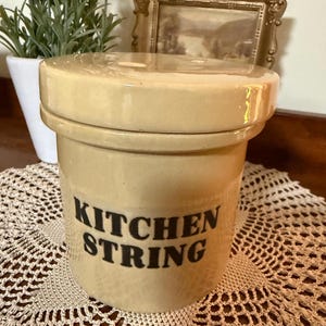 May include: A beige ceramic kitchen string holder with a matching lid. The words "KITCHEN STRING" are printed in black on the front. The holder sits on a cream-coloured, crocheted doily.