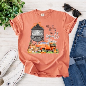 May include: A coral-colored t-shirt featuring a graphic of a water tower labeled 28320 BLADENBORO, NC, an orange pickup truck, and a pile of pumpkins. The text reads FALL IS BETTER IN A SMALL TOWN in black and script lettering on the front.
