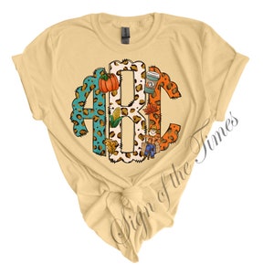 May include: A light yellow t-shirt with a monogram design featuring the letters A, B, and C. The letters are made of a leopard print pattern and are decorated with fall-themed elements like pumpkins, corn, and a scarecrow.