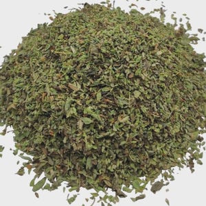May include: A mound of dried, crushed green herbs with varying shades of olive, forest green, and brown flecks. The texture is coarse and flaky, consisting of small, fragmented leaves and stems piled into a rounded, irregular heap against a plain background.