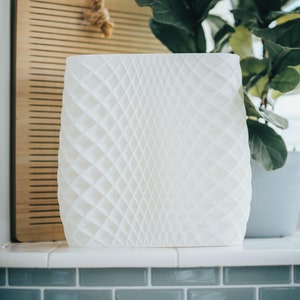 May include: White ceramic planter with a geometric diamond pattern. The planter is sitting on a tiled surface with a wooden cutting board and a green plant in the background.