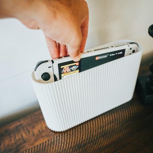 May include: A white, ribbed container holding a white and black handheld gaming console. The console's screen displays a game. The container sits on a dark wooden surface.