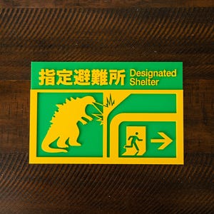 Può includere: Un cartello verde e giallo con testo giapponese e le parole inglesi "Designated Shelter". Il cartello presenta una silhouette di dinosauro gialla, fulmini e una freccia che indica una via di fuga.