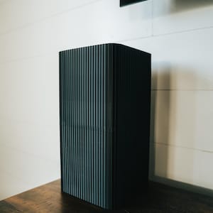 Könnte beinhalten: Ein hoher, rechteckiger schwarzer Lautsprecher mit vertikalen Rillen. Er steht auf einer dunklen Holzoberfläche vor einer weißen Wand. Der Lautsprecher hat ein modernes, minimalistisches Design.