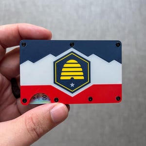 Puede incluir: Una cartera rectangular con la parte superior azul marino, el centro blanco y la parte inferior roja. La cartera presenta un emblema dorado y negro con una colmena y una estrella. Tornillos negros aseguran los componentes de la cartera.