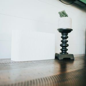 May include: A white rectangular object with a vertical ribbed texture sits next to a black pedestal candle holder. The candle holder features a stacked spherical design and supports a small white cylindrical planter containing a green succulent.