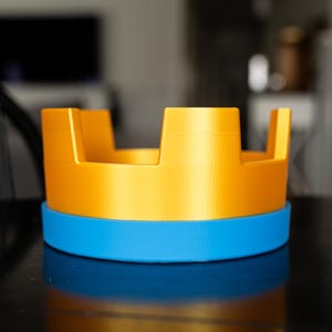 May include: A two-tone, 3D-printed object with a bright yellow top section and a blue base. The yellow portion has a crown-like design with rectangular cutouts. The object sits on a reflective black surface.