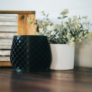 Pode incluir: Um vaso preto com um padrão geométrico de diamantes na sua superfície exterior. O vaso tem uma forma arredondada e bulbosa com uma base plana, apresentando um design de grelha consistente que cria uma aparência texturizada na parede.