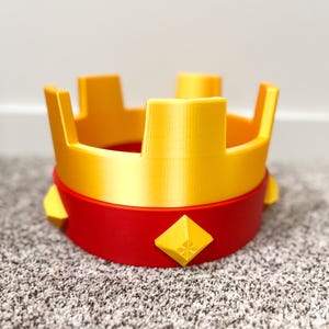 May include: A 3D-printed crown in yellow and red. The crown features a yellow upper section with four upright rectangular shapes. The lower section is red with yellow diamond-shaped accents. The crown sits on a carpeted surface.