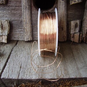 May include: A spool of copper wire sits on a weathered wooden surface. The wire is a warm, reddish-brown color, and some of it is unwound, creating loose loops. The background features rustic wooden elements.