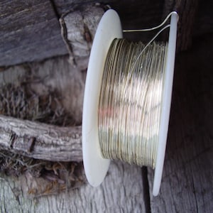 May include: A spool of gold-colored wire, tightly wound on a white plastic reel. The wire is thin and shiny, reflecting light. The reel is positioned against a weathered wooden background, suggesting a craft or jewelry-making theme.