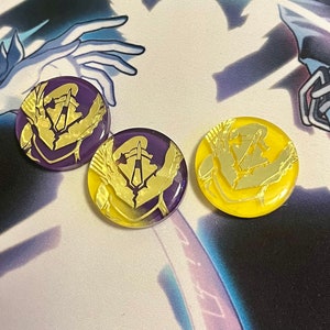 May include: Three round, decorative pins with a gold design on a purple and yellow background. The design features a stylized, geometric shape with a central point.