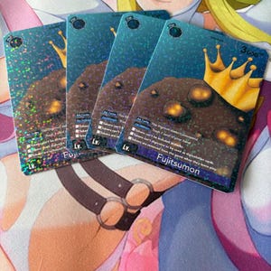 May include: Four trading cards featuring a brown creature with a golden crown, set against a blue, starry background. The cards have text including "Fujitsumon" and "3600". The cards are on a colorful surface.