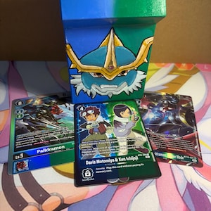 May include: A hand-painted, square, blue and green container with a cartoon character design, next to three Digimon trading cards. The cards feature characters and text, including "Paildramon" and "Davis Motomiya & Ken Ichijoji".