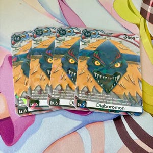 Pode incluir: Várias cartas colecionáveis ​​Digimon com Diaboromon. As cartas apresentam uma ilustração de monstro azul e laranja, o número 14 e "Lv.6". As cartas estão empilhadas em uma superfície rosa e branca.