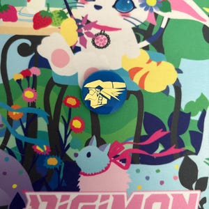 Könnte beinhalten: Ein blauer Digimon-Kartenspiel-Pin mit einem goldenen Charakterdesign. Der Pin befindet sich vor einem farbenfrohen Hintergrund mit einem weißen Kaninchen mit floralen Akzenten, Erdbeeren und dem Text "Digimon Card Game".