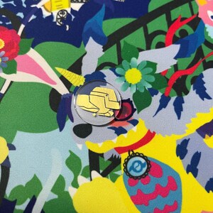 May include: A colorful fabric with a whimsical design featuring a yellow and white unicorn, a yellow and pink flower, and a yellow and blue bird. A small gold pin with a yellow hat is attached to the fabric.