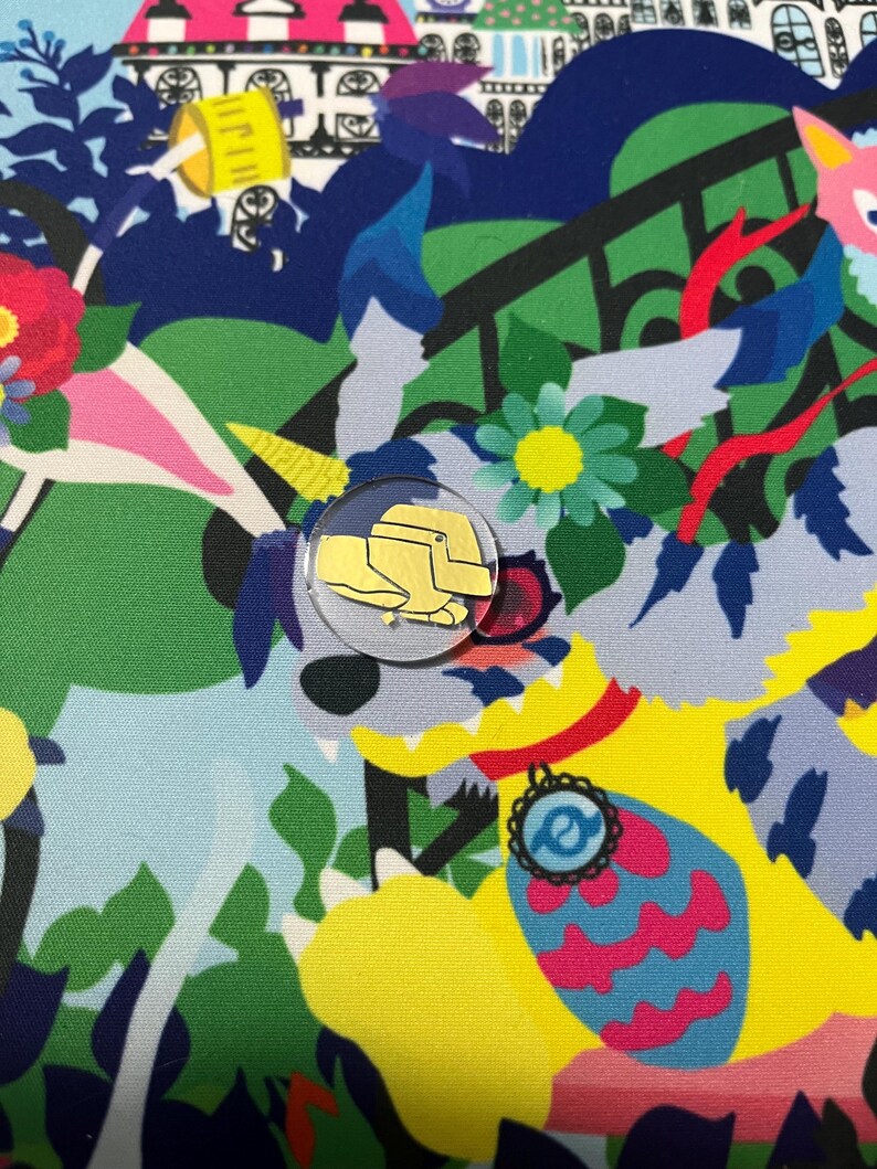 May include: A colorful fabric with a whimsical design featuring a yellow and white unicorn, a yellow and pink flower, and a yellow and blue bird. A small gold pin with a yellow hat is attached to the fabric.