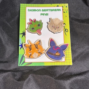 May include: Four wooden pins featuring stylised character faces attached to a rectangular display card. The card has a lime green border with a grid pattern and the text DIGIMON BEATBREAK PINS! The pins depict a green creature, a tan lion-like face, an orange striped cat, and a blue masked character.