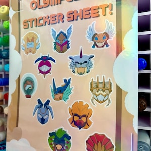 May include: A rectangular sticker sheet titled OLYMPUS 12 STICKER SHEET! featuring twelve distinct, colourful character head illustrations. The designs include various mythical and creature-like figures with diverse shapes, colours, and headgear.
