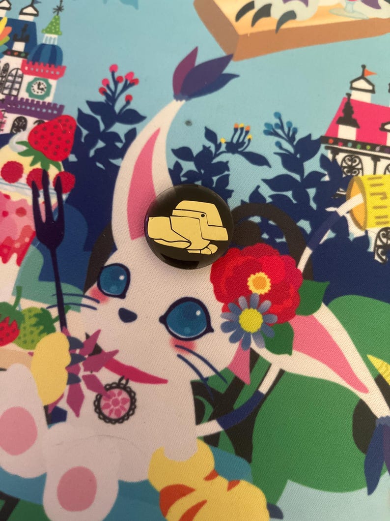 May include: A black and gold button pin with a cartoon image of a yellow duck wearing a hat. The pin is on a colorful background with a white cat with blue eyes.