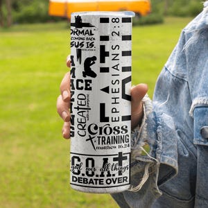 May include: A white tumbler with black text and graphics. The tumbler features phrases like "NORMAL COMING BACK SUS IS," "EPHESIANS 2:8," and "Cross TRAINING." The design includes a silhouette of a person kneeling and a cross. The tumbler is held by a person.
