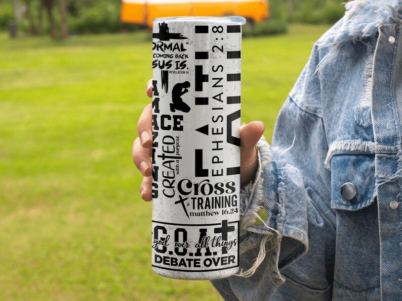 May include: A white tumbler with black text and graphics. The tumbler features phrases like "NORMAL COMING BACK SUS IS," "EPHESIANS 2:8," and "Cross TRAINING." The design includes a silhouette of a person kneeling and a cross. The tumbler is held by a person.