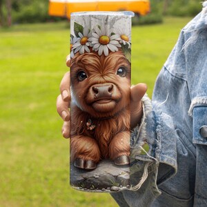 May include: A stainless steel tumbler featuring a detailed illustration of a Highland calf with a daisy flower crown. The calf is brown with large eyes and a small butterfly. The tumbler is held against a green background.